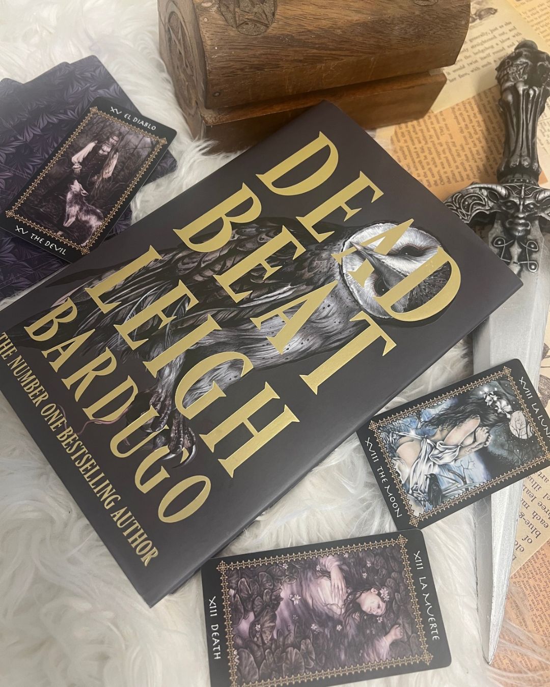 Dead Beat by Leigh Bardugo, featuring its black and gold owl cover surrounded by Gothic tarot cards, an ornate dagger and occult accessories in a dark academia books flat lay.
